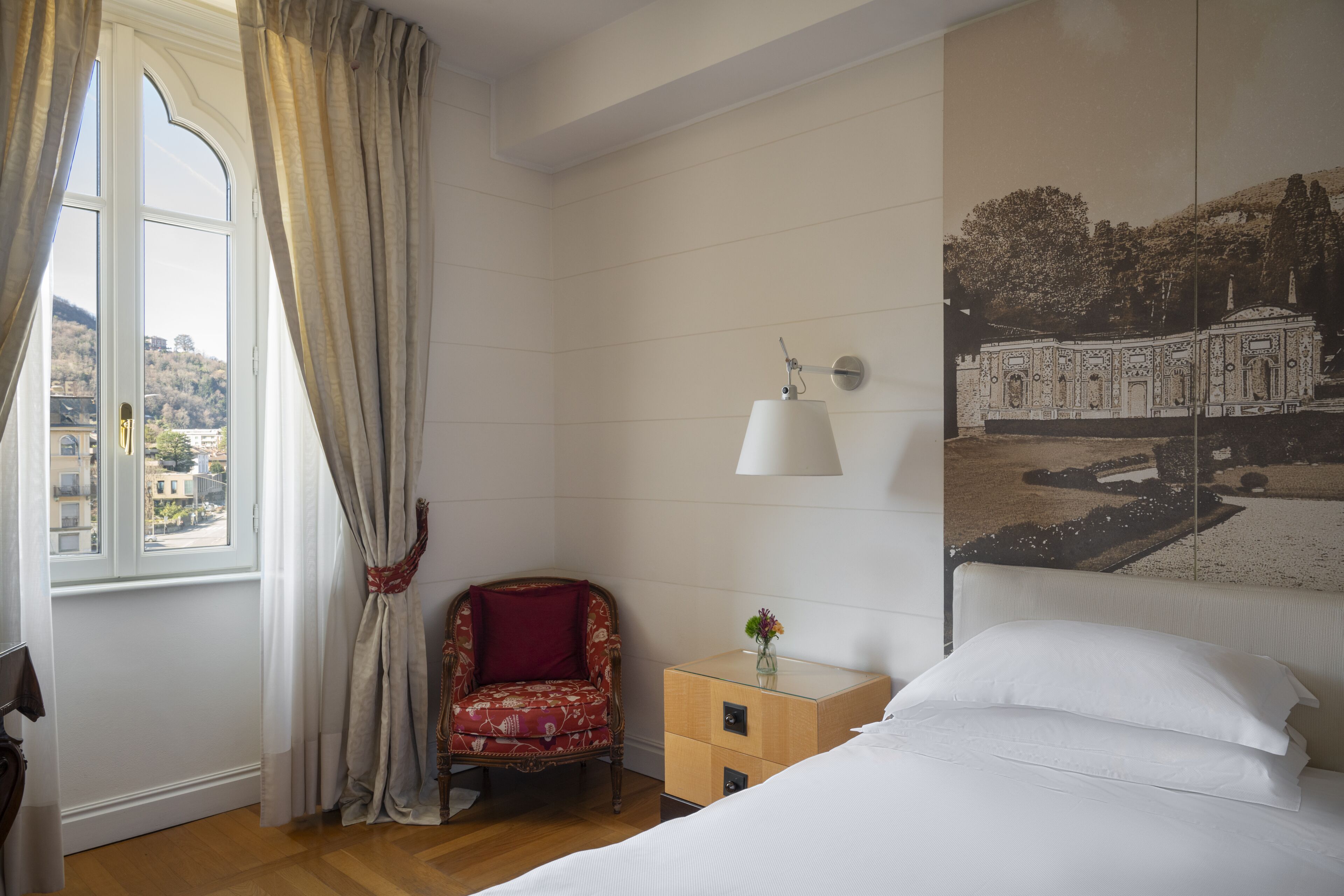 A hotel room with a bed, a chair, a nightstand, a lamp, and a window with curtains.