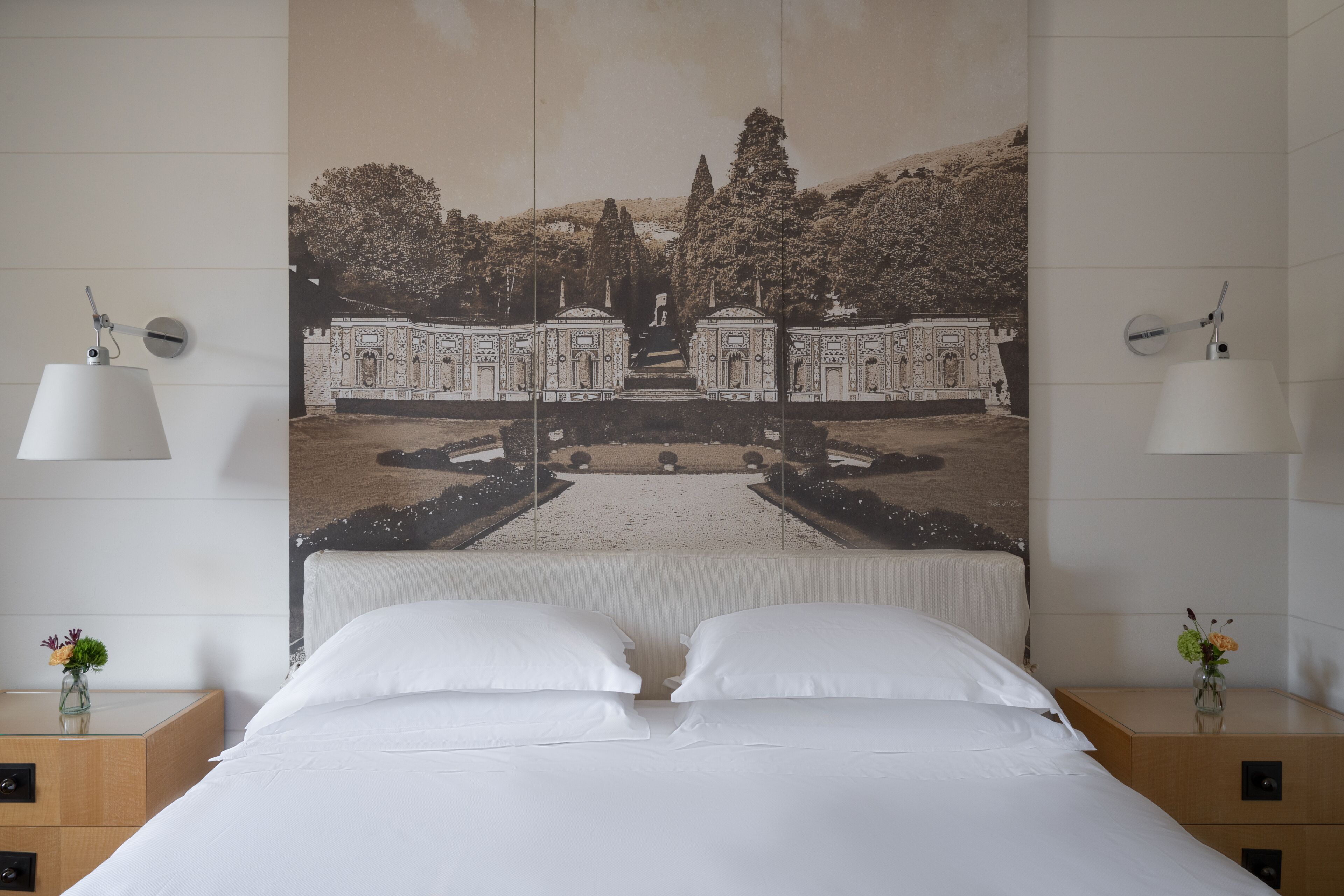 Una camera d'albergo con un letto grande, due comodini con lampade, uno specchio fissato al muro e un quadro incorniciato di un giardino.