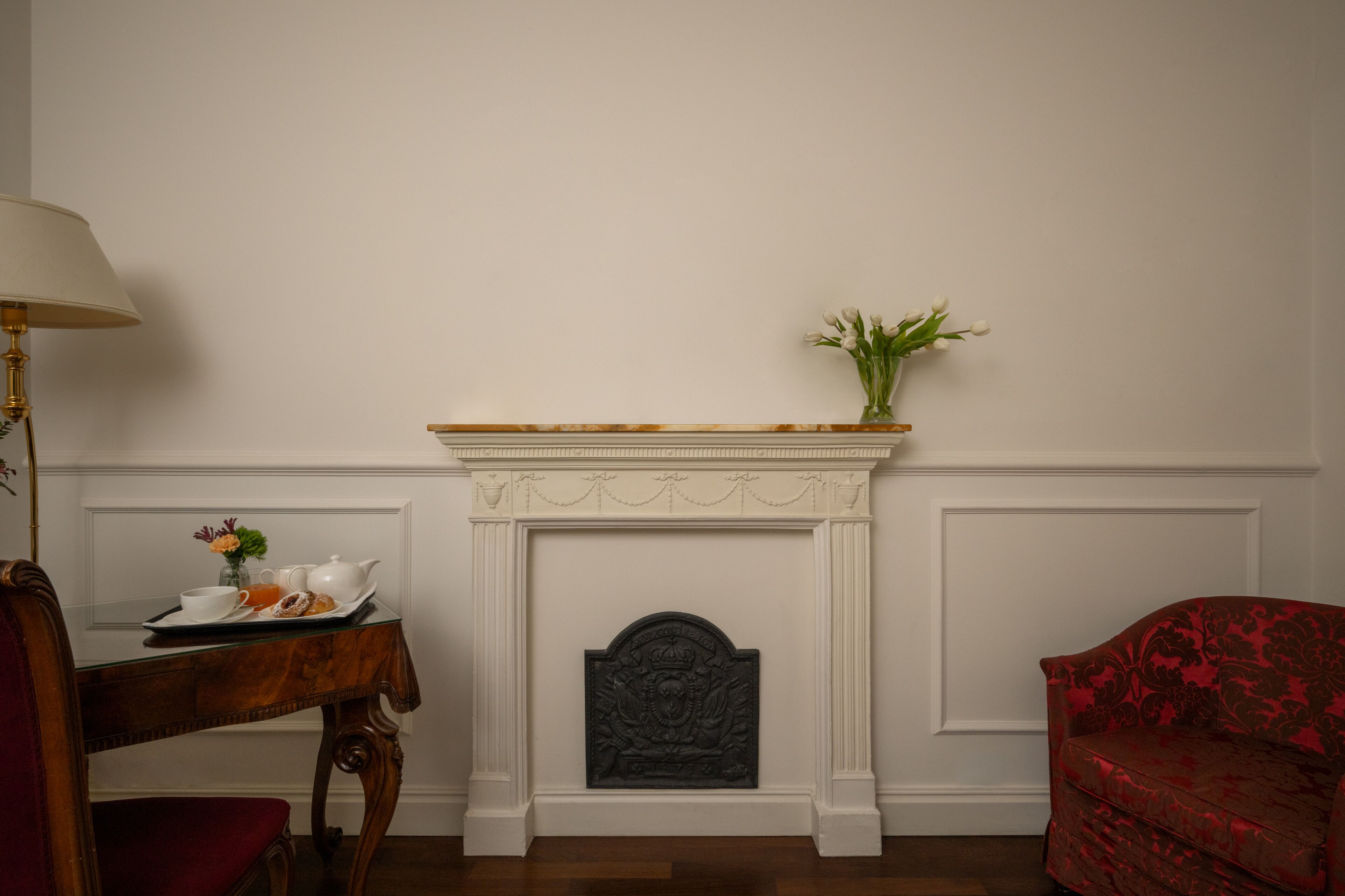 A room with a fireplace, a table with a tray of breakfast, a red chair, and a lamp.