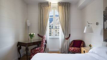 Quarto de hotel com cama, escrivaninha, duas cadeiras, janela com cortinas e luminária.
