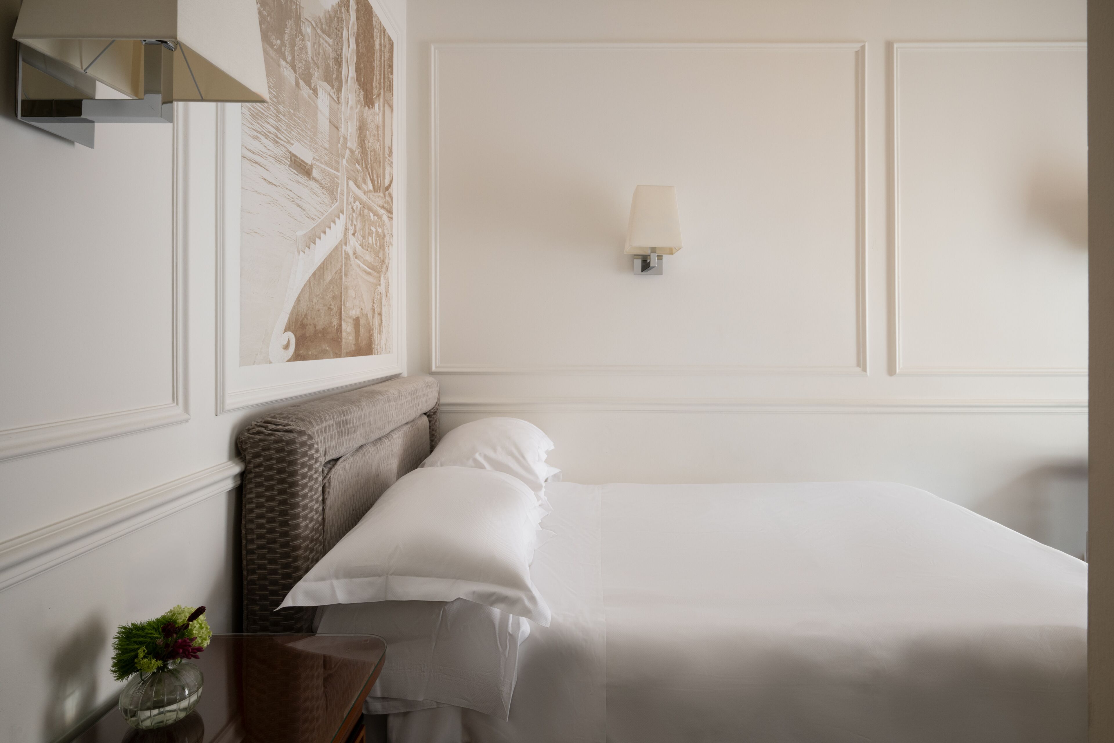 Una cama bien hecha en una habitación de hotel, con una lámpara de pared, un adorno mural y una mesita con un florero.