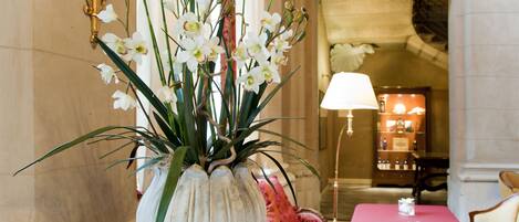 Una habitación con piso de mármol, una mesa con mantel rojo, un jarrón con flores blancas y una lámpara.