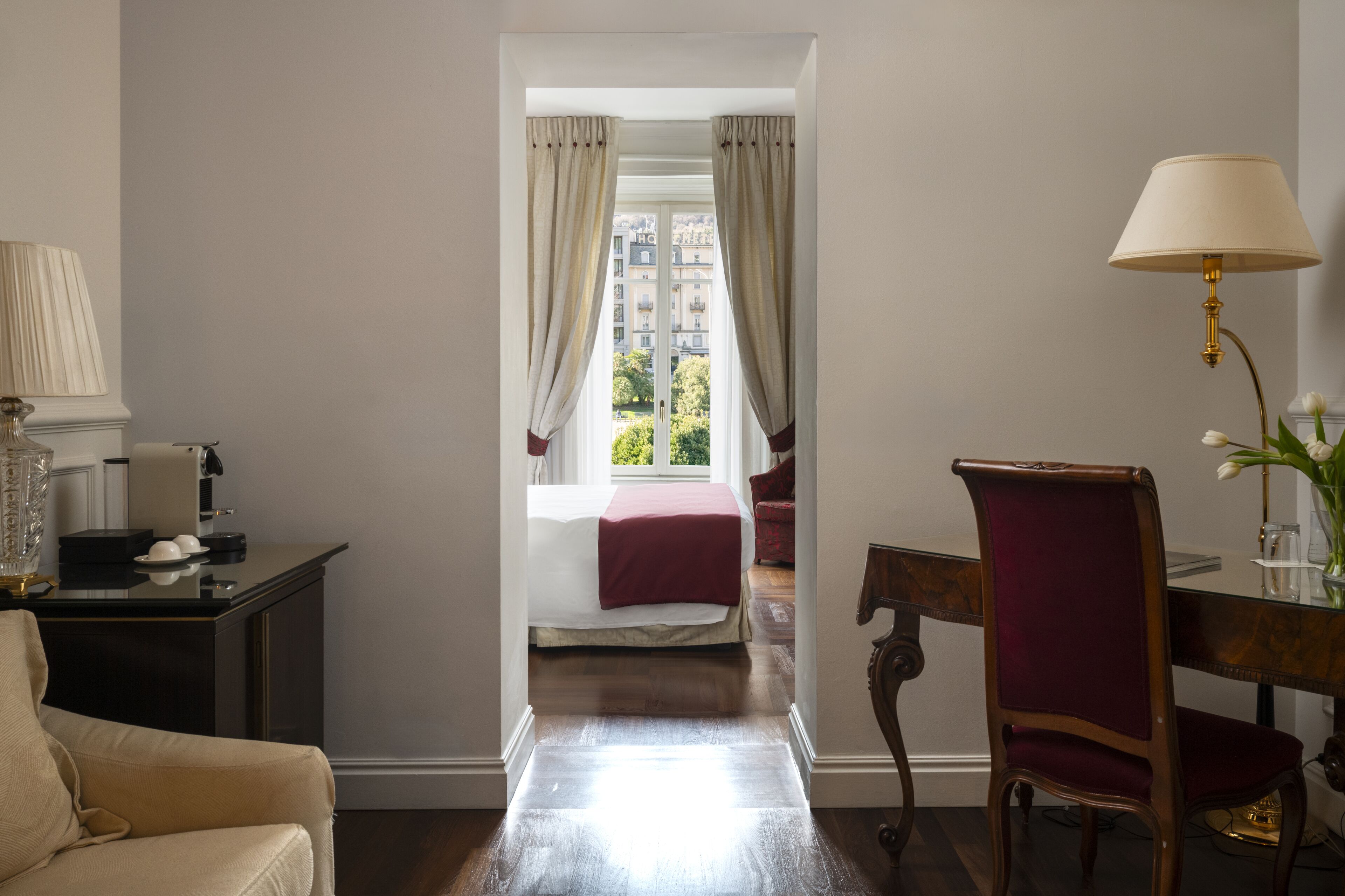 A hotel room with a bed, a desk, a chair, a lamp, and a view of buildings through the window.