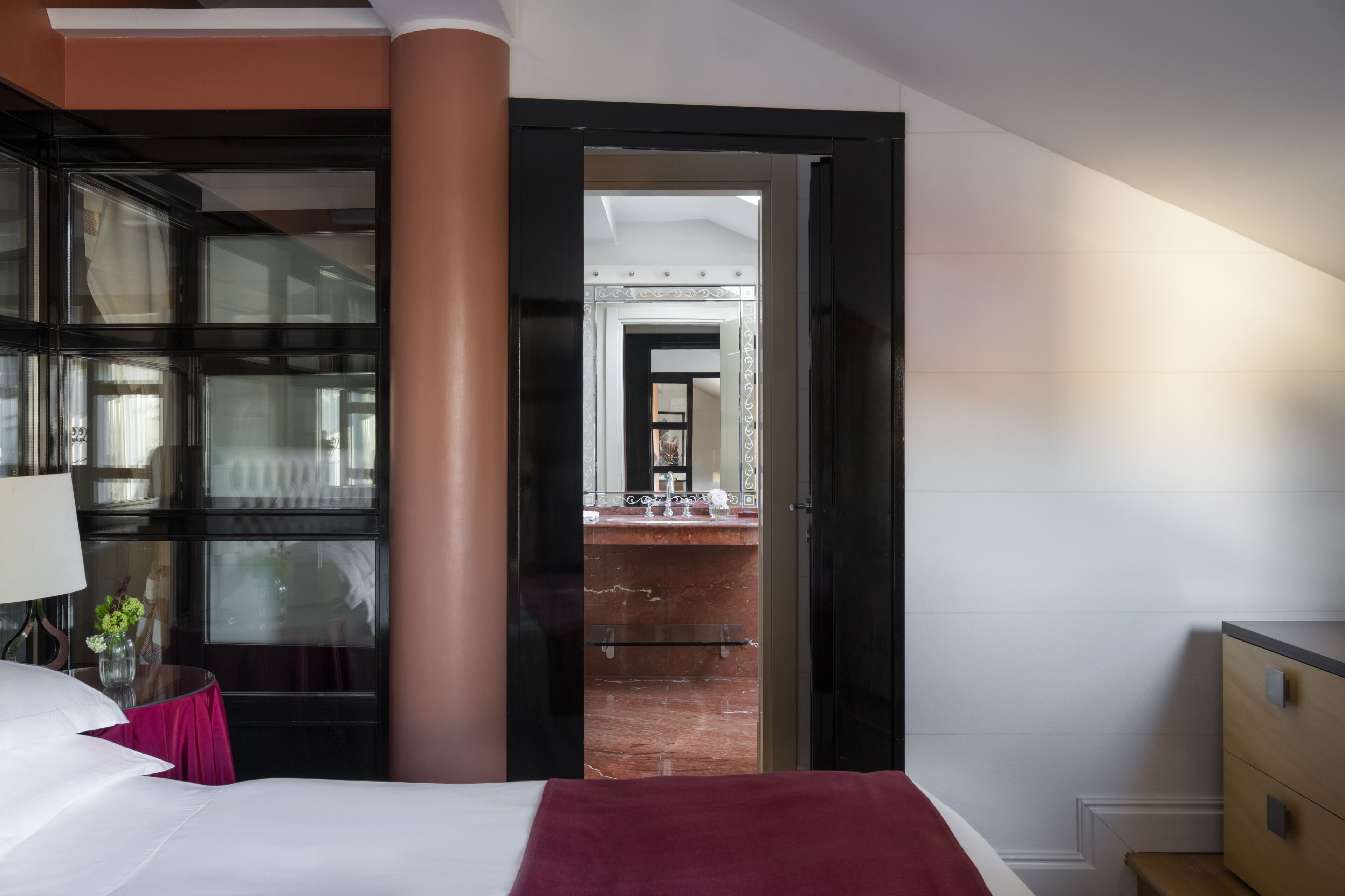 Camera d'albergo con un letto, una scrivania con lampada, un vaso di fiori e vista sul bagno attraverso una porta aperta.