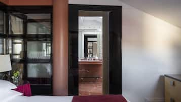 Habitación de hotel con cama, escritorio con lámpara, un florero y vista al baño por una puerta abierta.