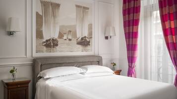 Un lit bien fait, avec du linge de lit blanc, un grand tableau encadré représentant des voiliers accroché au mur, et une fenêtre ornée de rideaux roses.