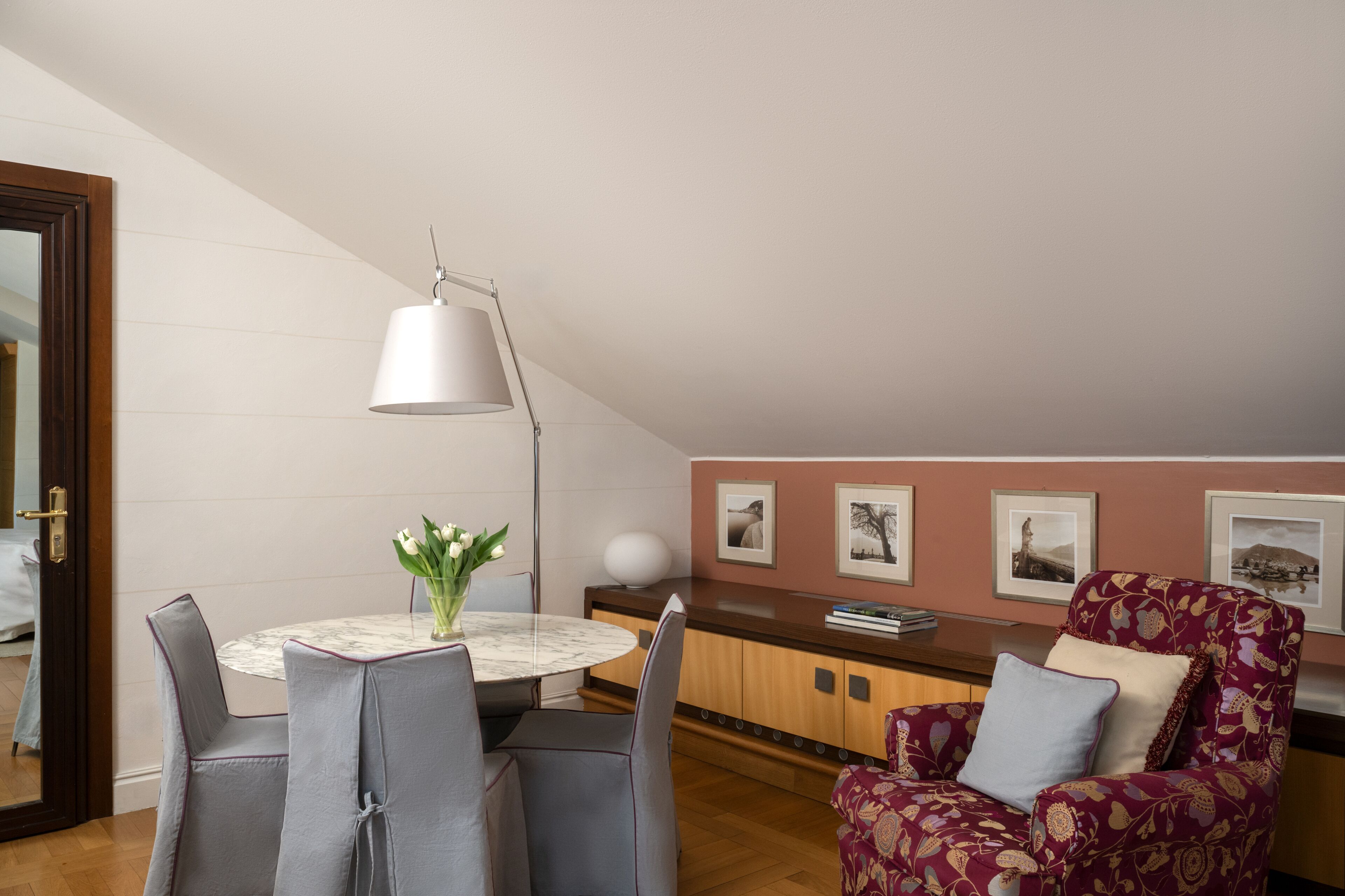 A dining area with a round table, chairs, a vase with flowers, a lamp, and framed pictures on the wall.