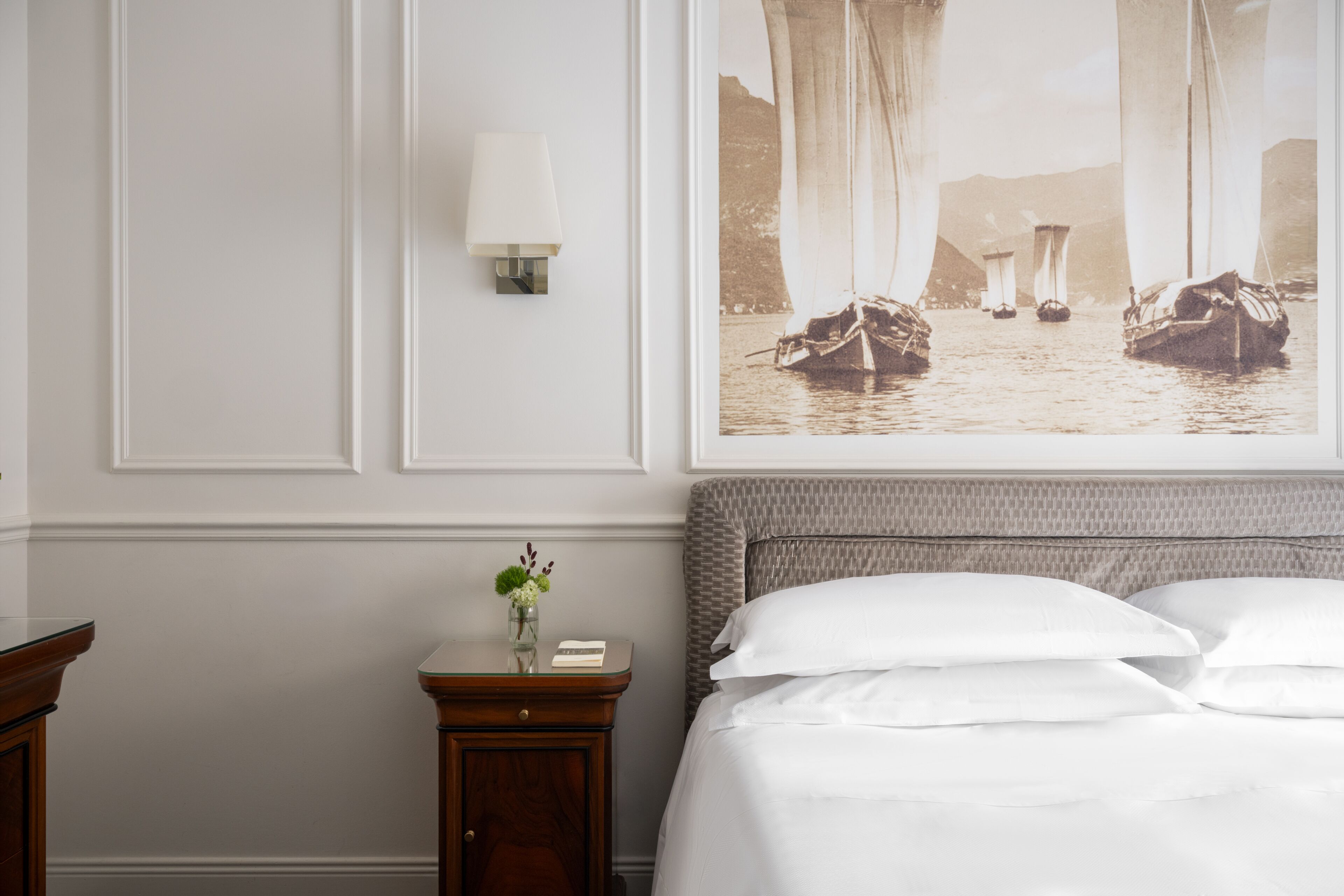 A hotel room with a bed, a nightstand with a vase of flowers, and a wall-mounted lamp.