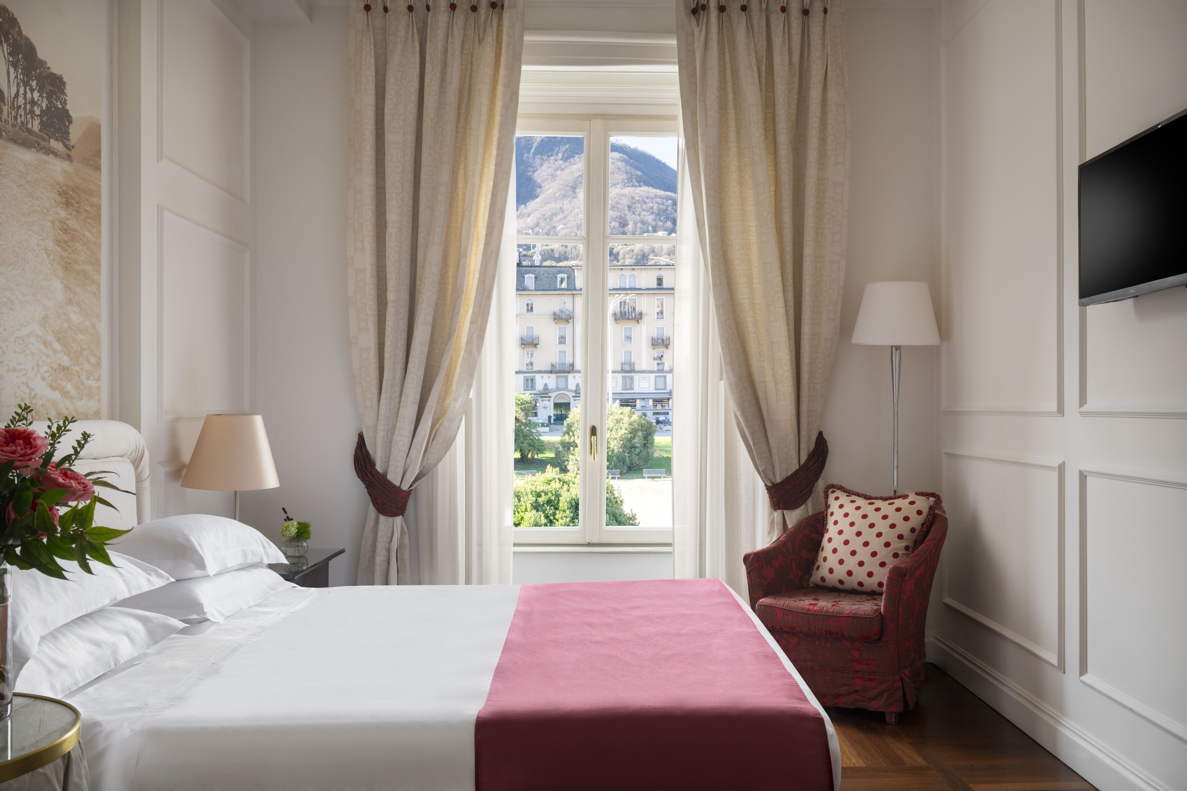 Una habitación de hotel con cama, una silla, televisor y ventana con cortinas.