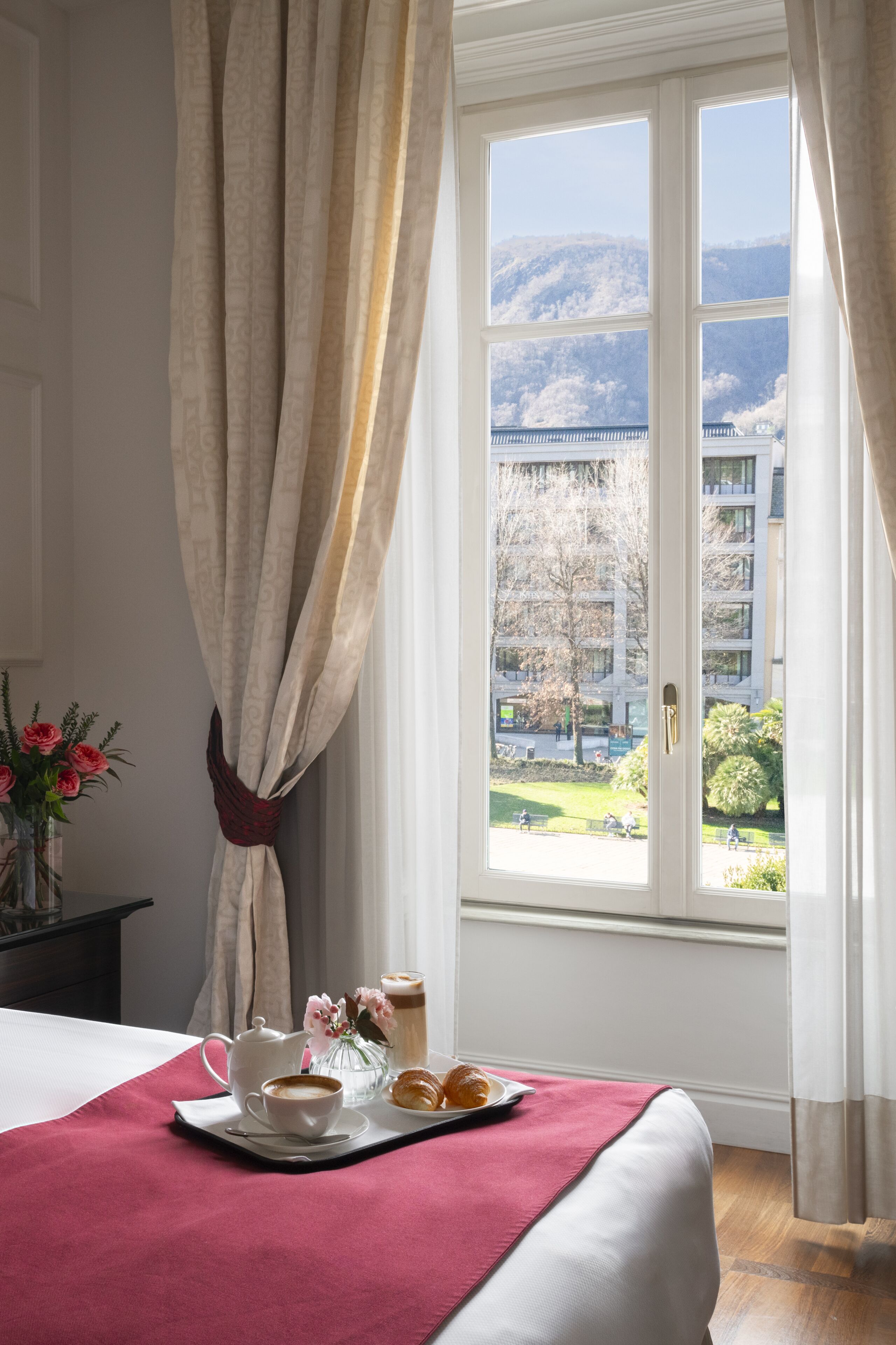 A hotel room with a bed, a tray with breakfast items, a vase with flowers, and a view of a park and buildings through a window.