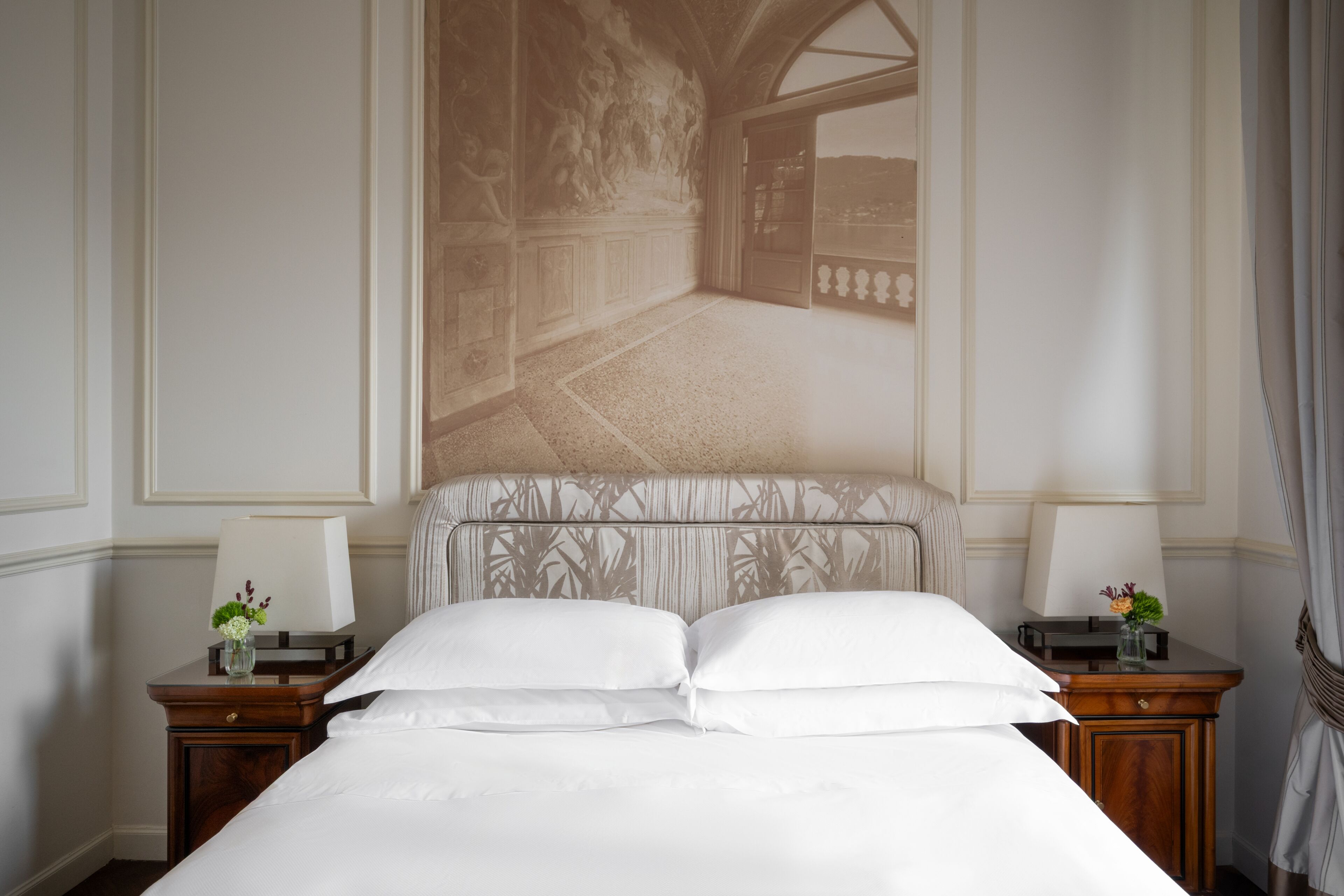 A hotel room with a large bed, two bedside tables, a nightstand, a wall-mounted lamp, and a view through a window.