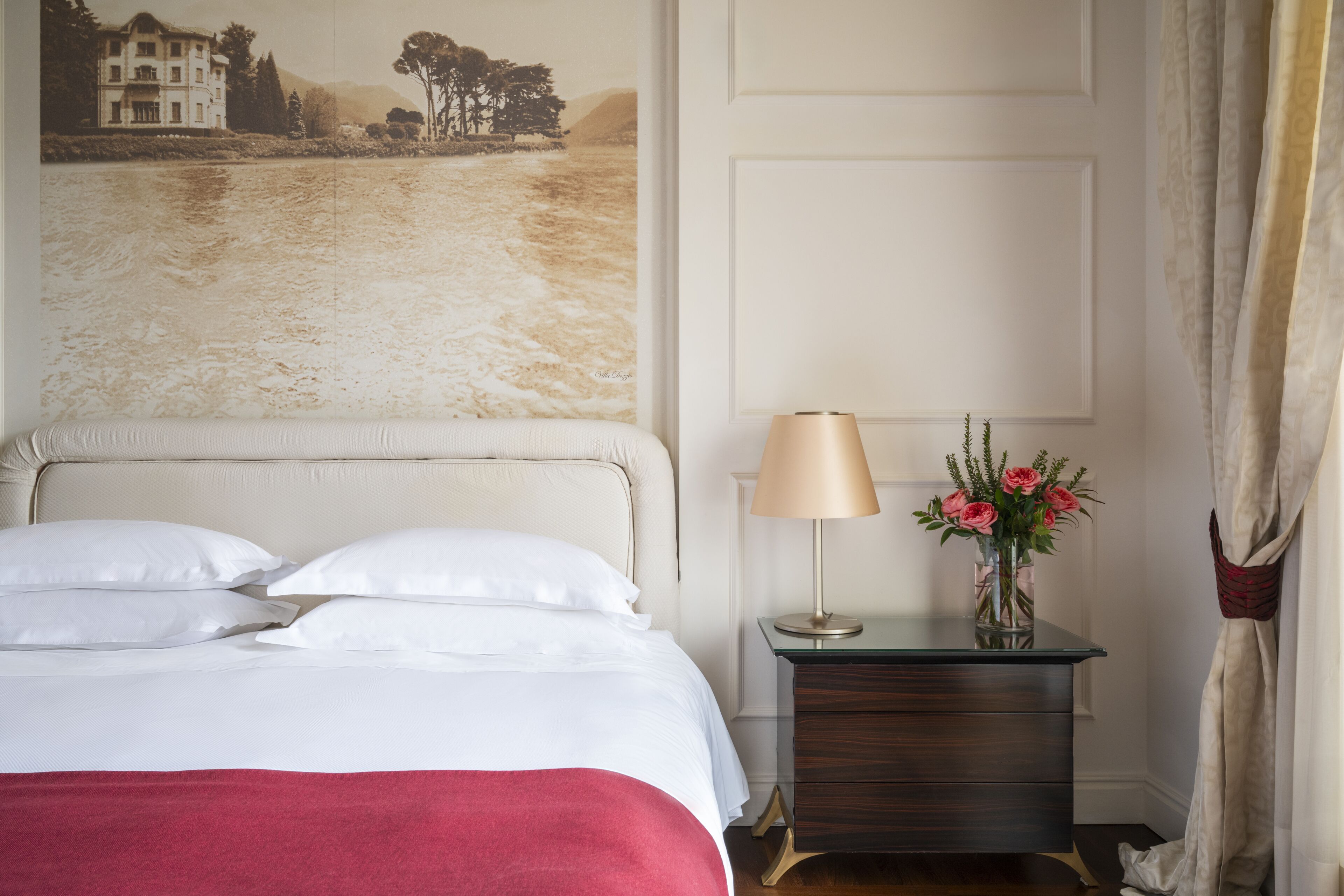 Une chambre d’hôtel avec un lit, une table de chevet avec une lampe et des fleurs, un mur orné d’un tableau et un rideau.