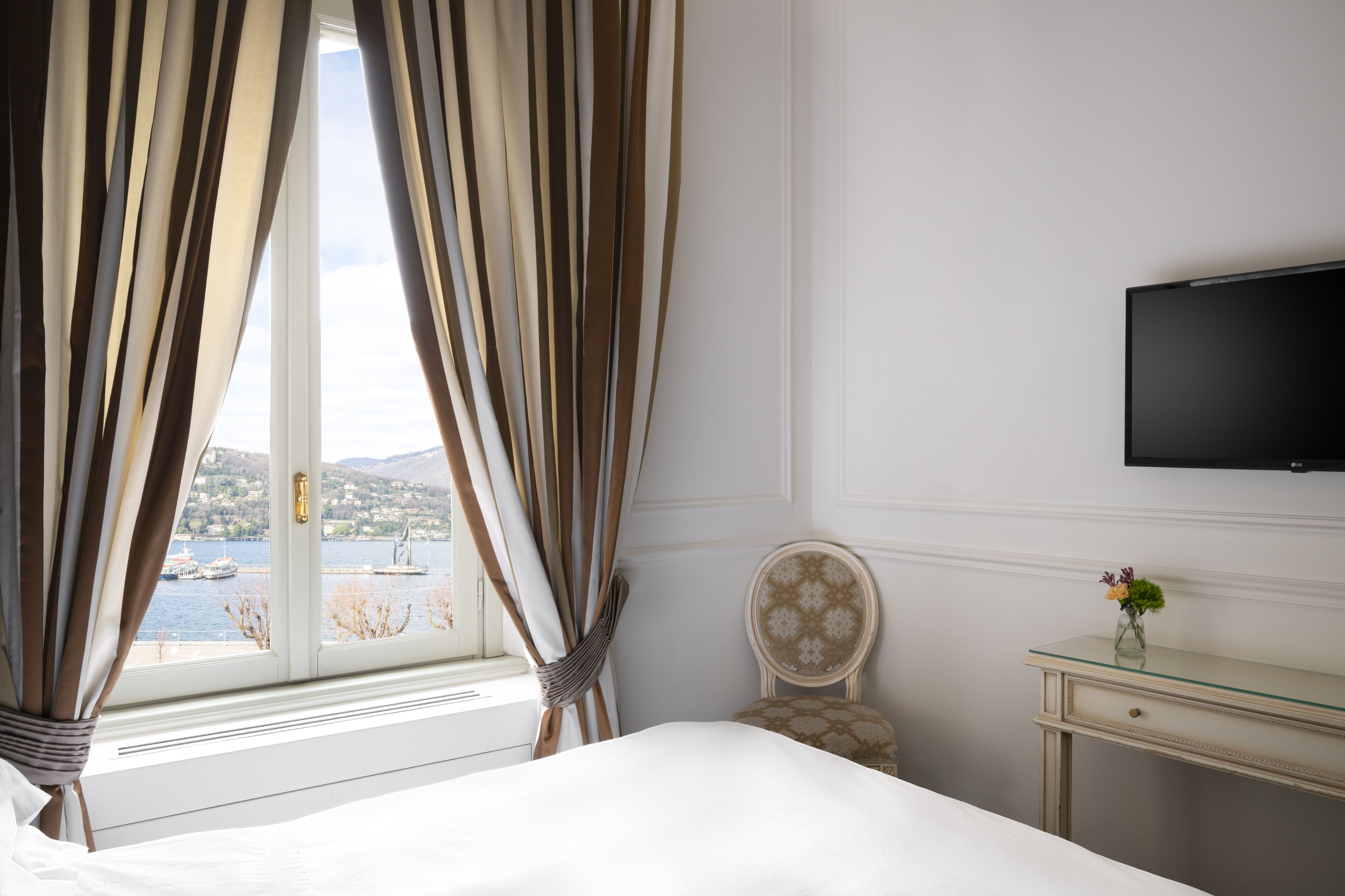 Una habitación de hotel con cama, una silla, un escritorio, televisión y ventana con cortinas.