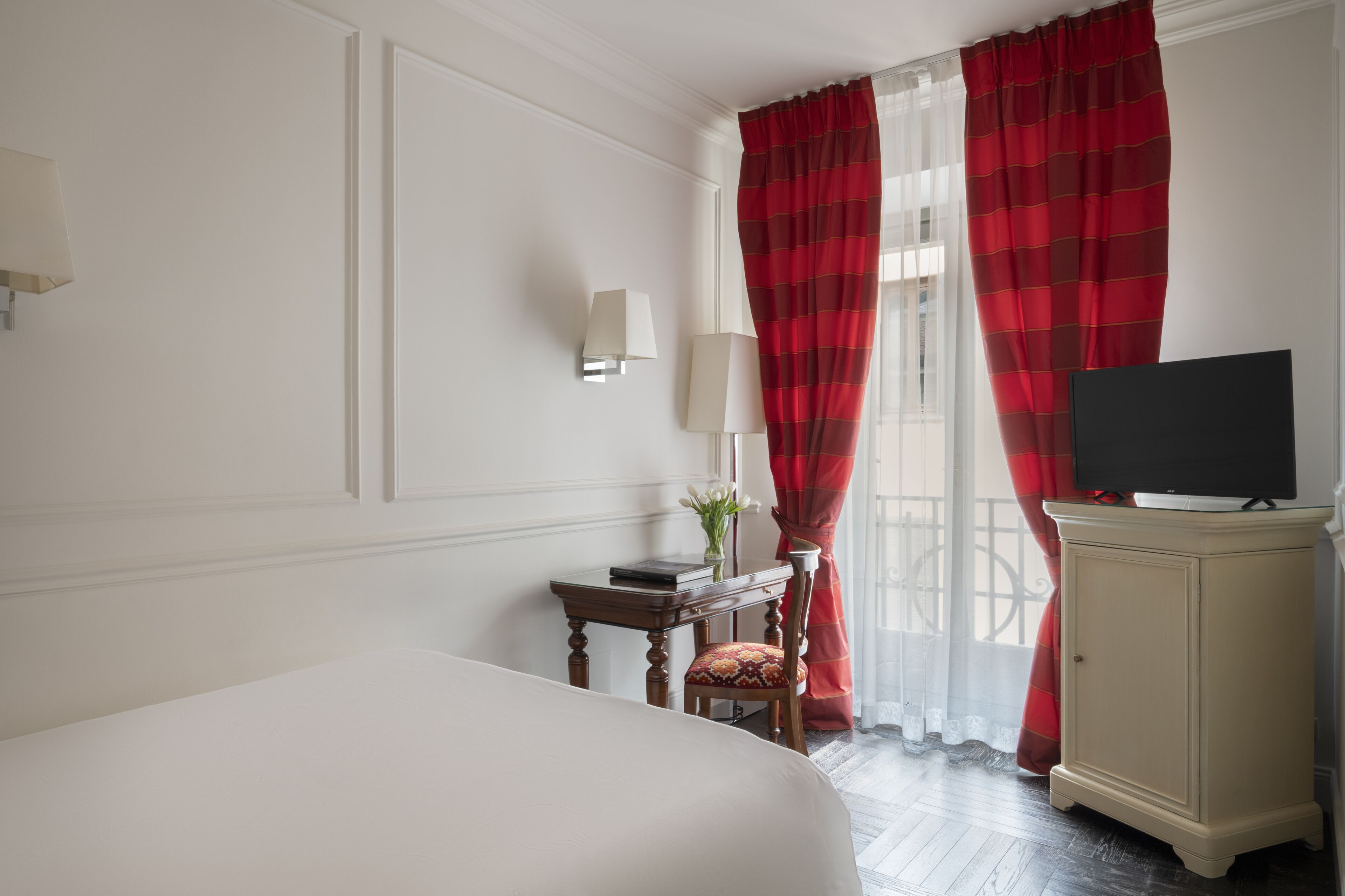 A hotel room with a bed, a desk with a lamp, a television on a cabinet, and red curtains.