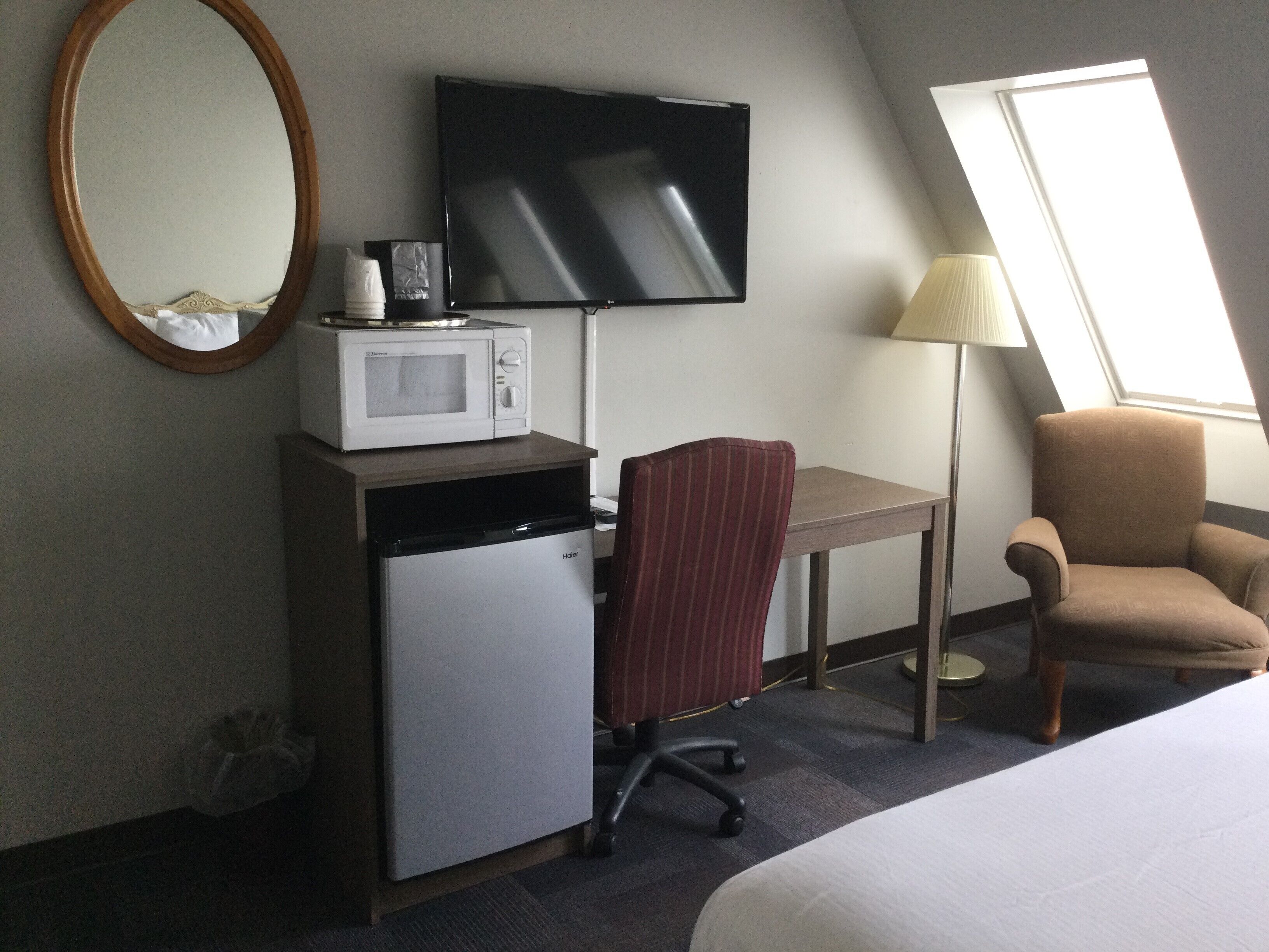 A hotel room with a bed, a desk with a microwave, a television, a chair, and a lamp.