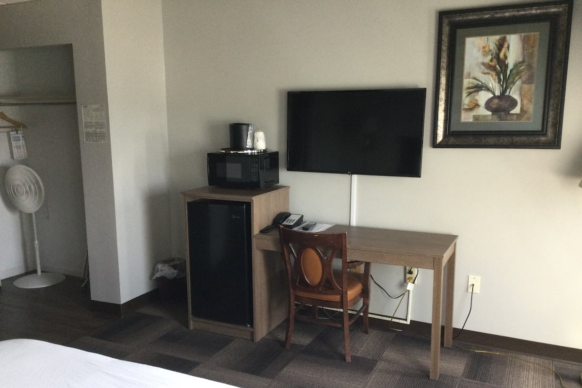 A hotel room with a wooden desk, a chair, a flat-screen TV mounted on the wall, a microwave on a cabinet, and a fan.