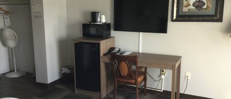 Una habitación de hotel con un escritorio de madera, una silla, un televisor de pantalla plana montado en la pared, un microondas en un mueble y un ventilador.