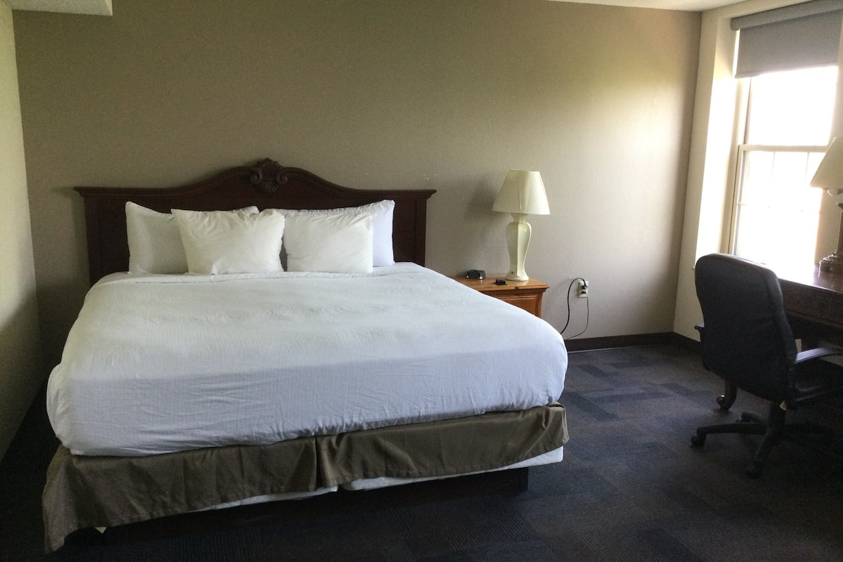 A neatly made bed with white linens, a wooden headboard, a bedside lamp, and a desk with a chair in a hotel room.