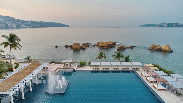 Piscina de resort com espreguiçadeiras e guarda-sóis, uma fonte de água e vista para o mar, com pedras e palmeiras.
