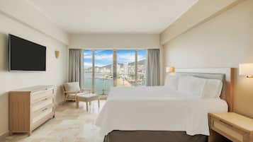 A hotel room with a large bed, a TV, a wooden dresser, a chair, and a view of a cityscape and water.