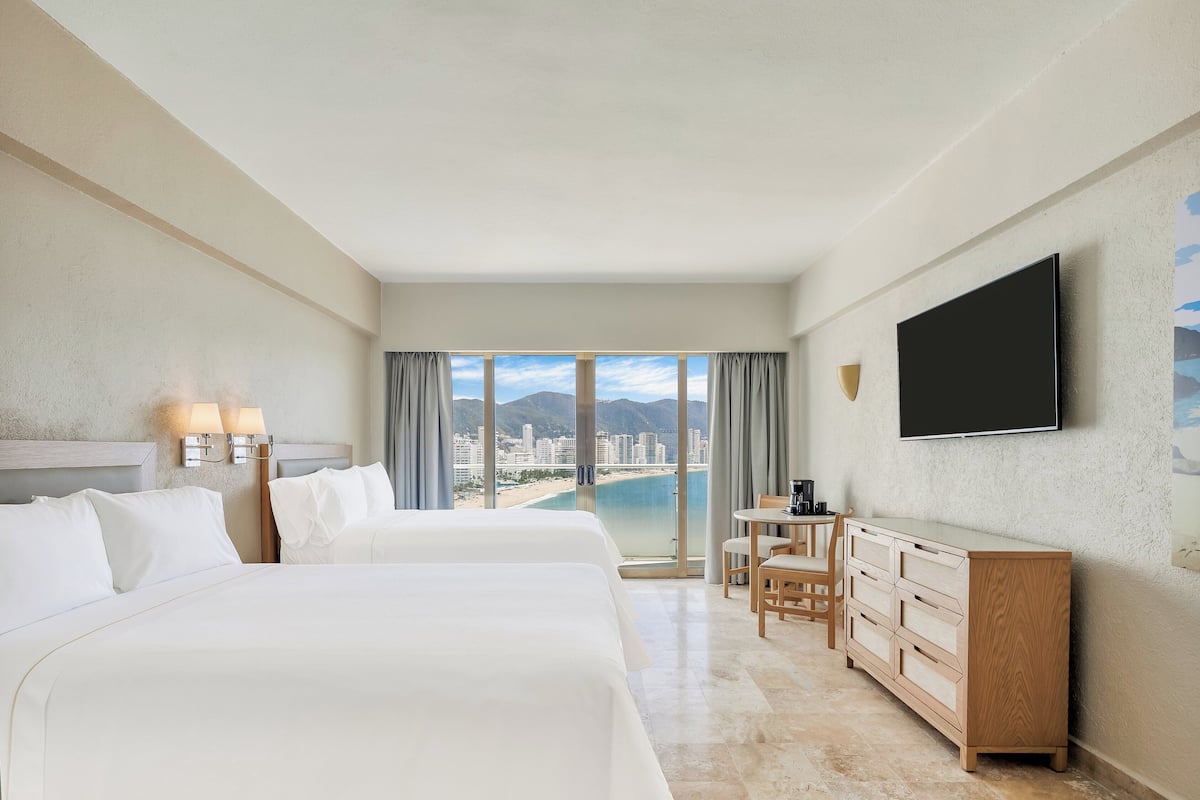 A hotel room with two beds, a TV, a desk, and a view of a city and beach.
