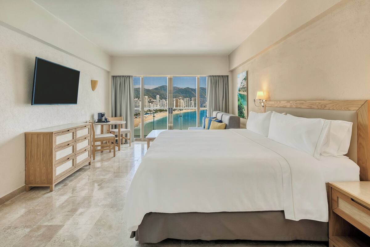 A hotel room with a large bed, a TV mounted on the wall, a desk with a chair, and a view of a beach and mountains through the windows.