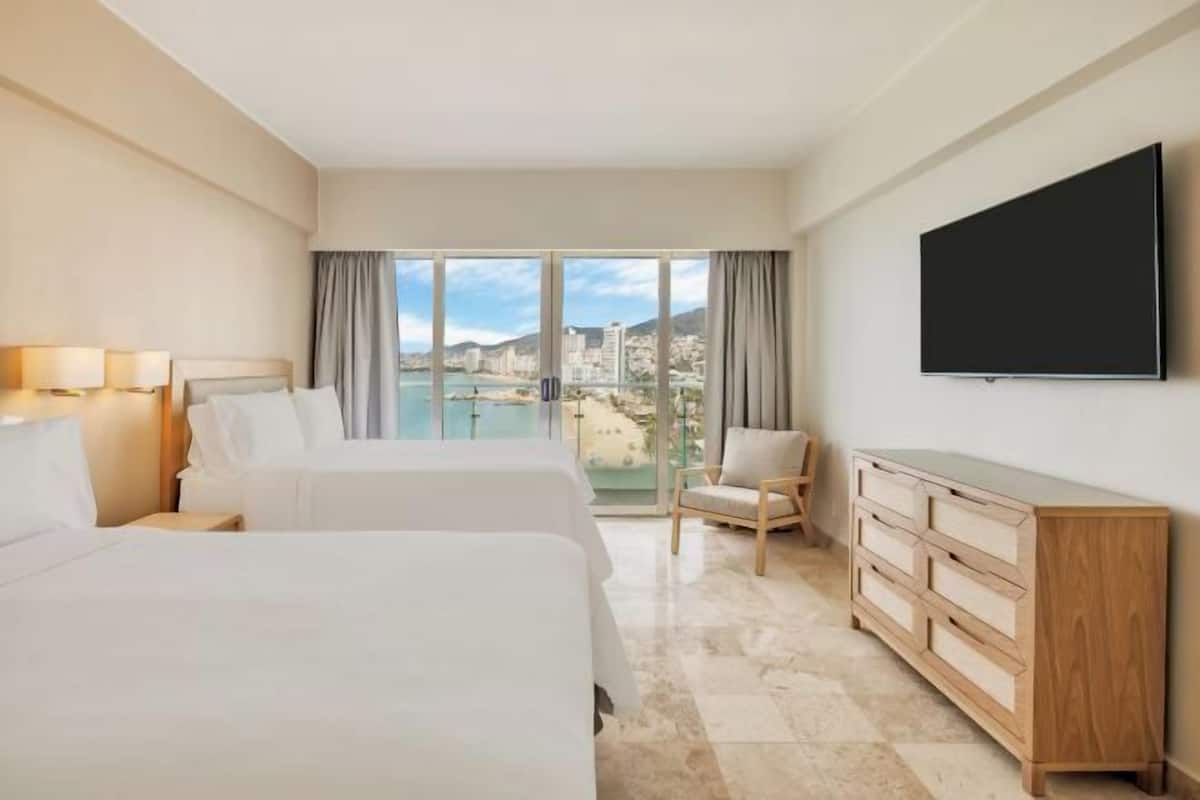 A hotel room with two beds, a TV, a chair, a dresser, and a large window with a city view.
