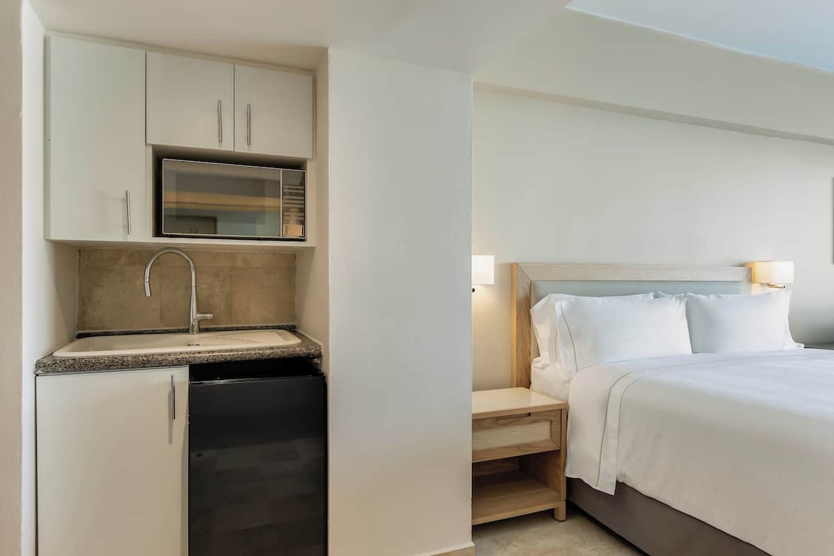 A compact hotel room with a kitchenette, a bed with white linens, and a nightstand with a lamp.