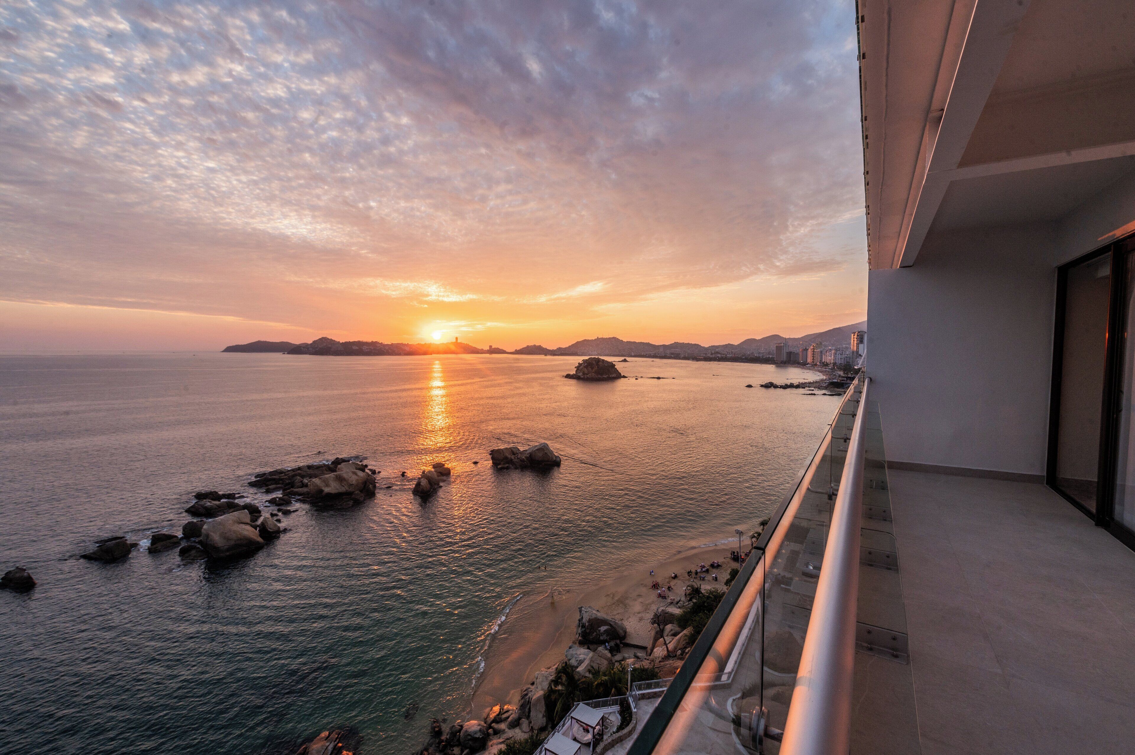 Un balcón con vista al mar al atardecer, con afloramientos rocosos y un horizonte urbano distante.