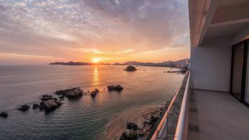Un balcón con vista al mar al atardecer, con afloramientos rocosos y un horizonte urbano distante.