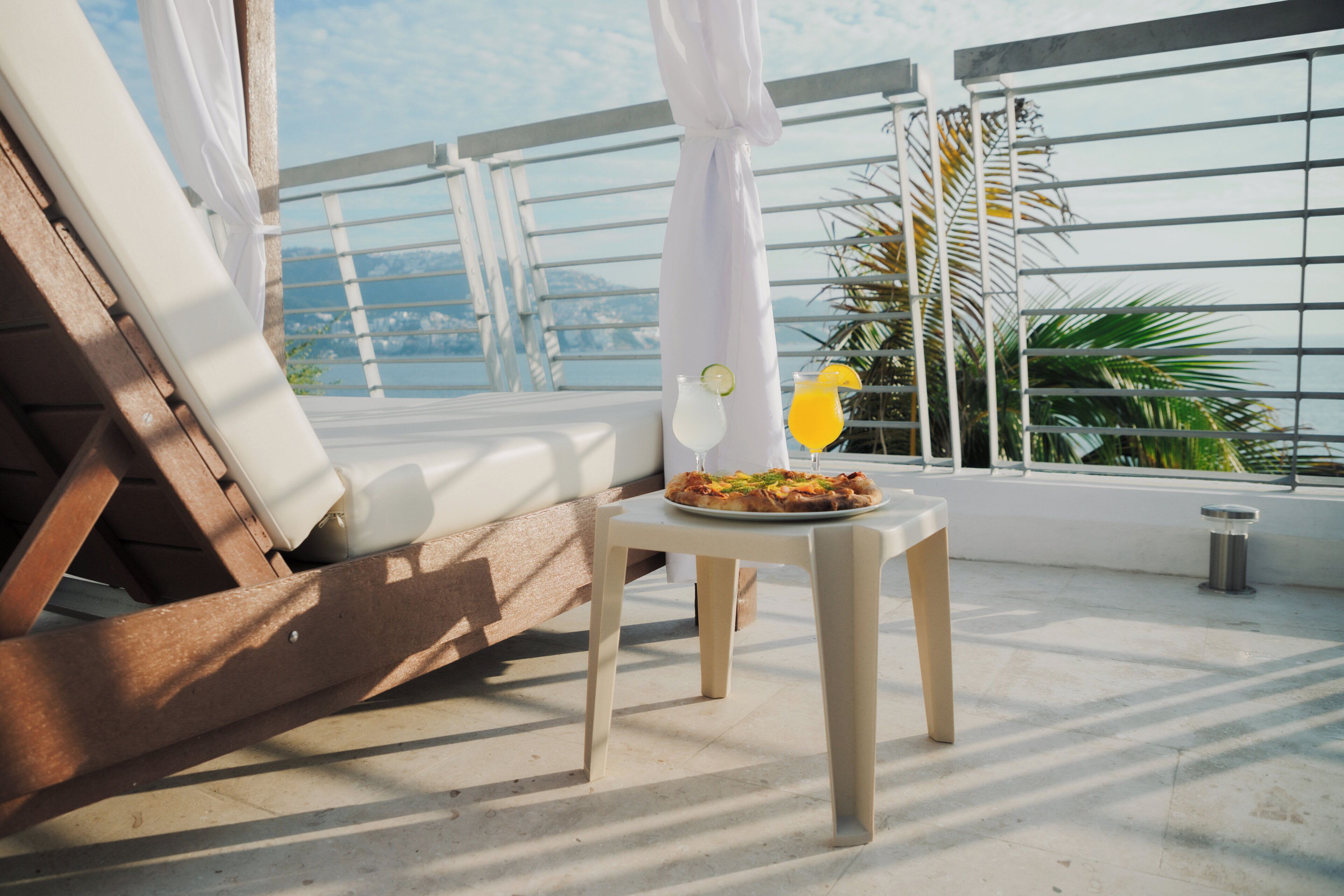 Una terraza iluminada por el sol con una mesa dispuesta para comer, una silla de descanso y vistas al mar y a las montañas.