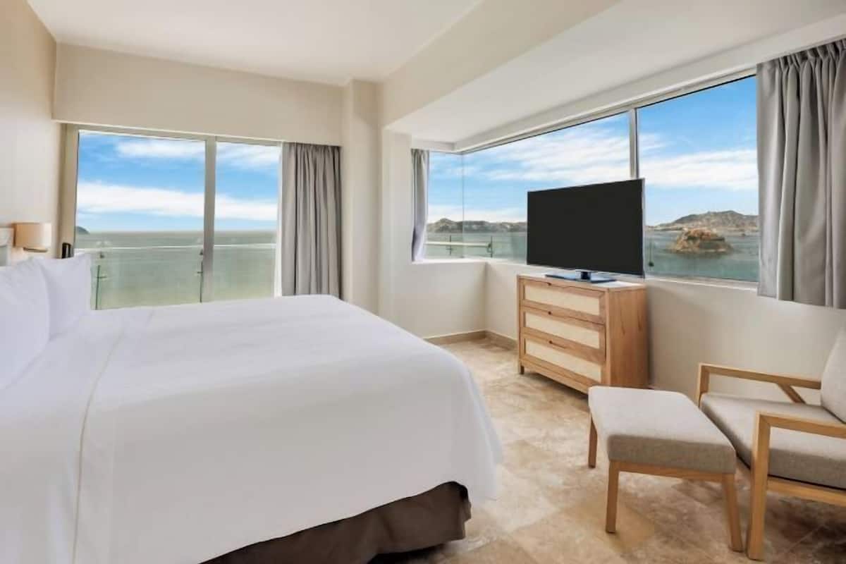 A hotel room with a large bed, a TV, a wooden dresser, and a view of the sea.