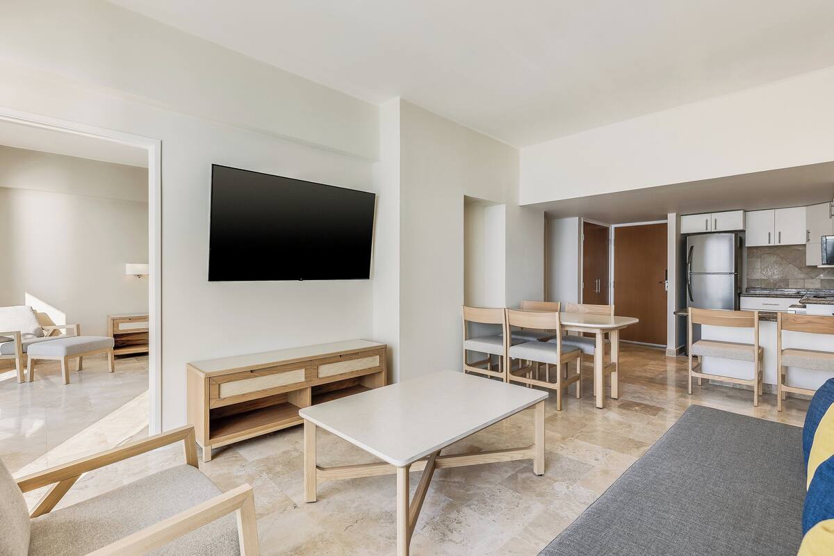 A modern living room with a flat-screen TV, wooden furniture, and a kitchen area with cabinets and a refrigerator.