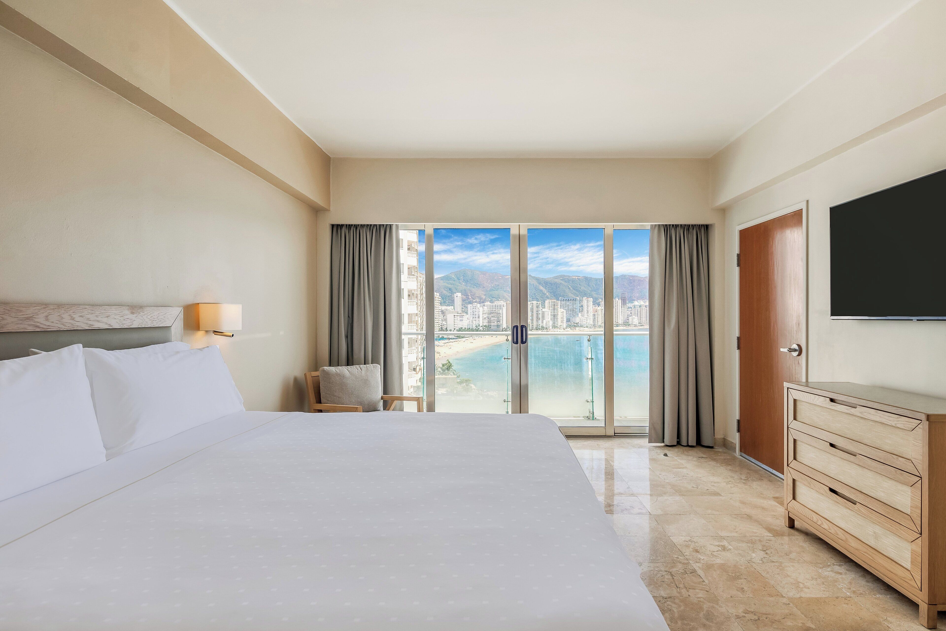 A hotel room with a large bed, a dresser, a TV, and a view of a beach and cityscape through large windows.