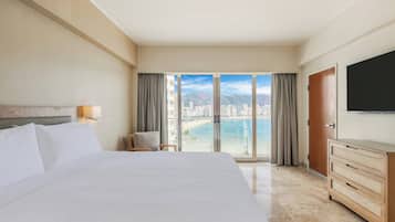 A hotel room with a large bed, a dresser, a TV, and a view of a beach and cityscape through large windows.