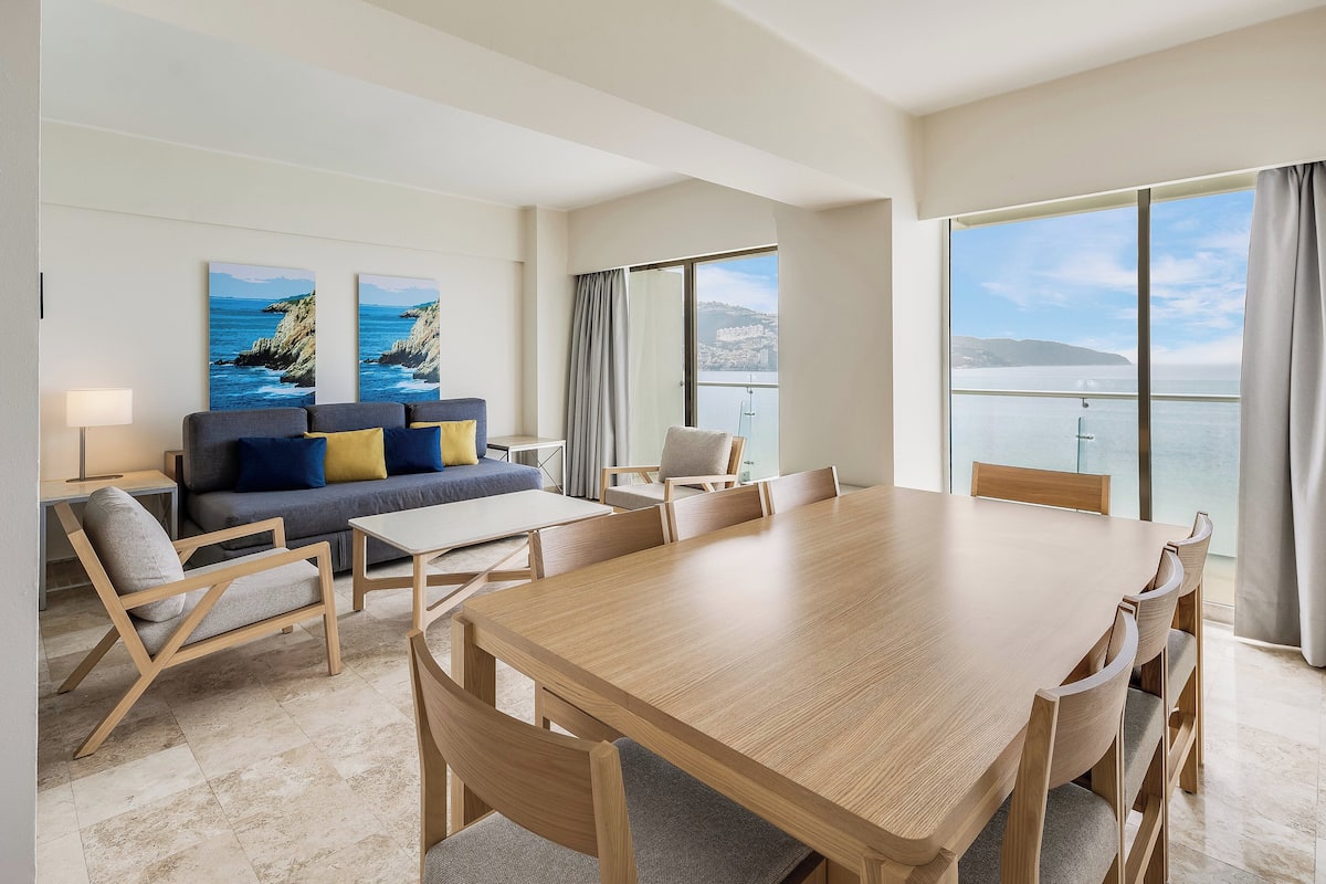 A modern hotel room with a large dining table, comfortable seating, a sofa, and a balcony with a view of the sea.