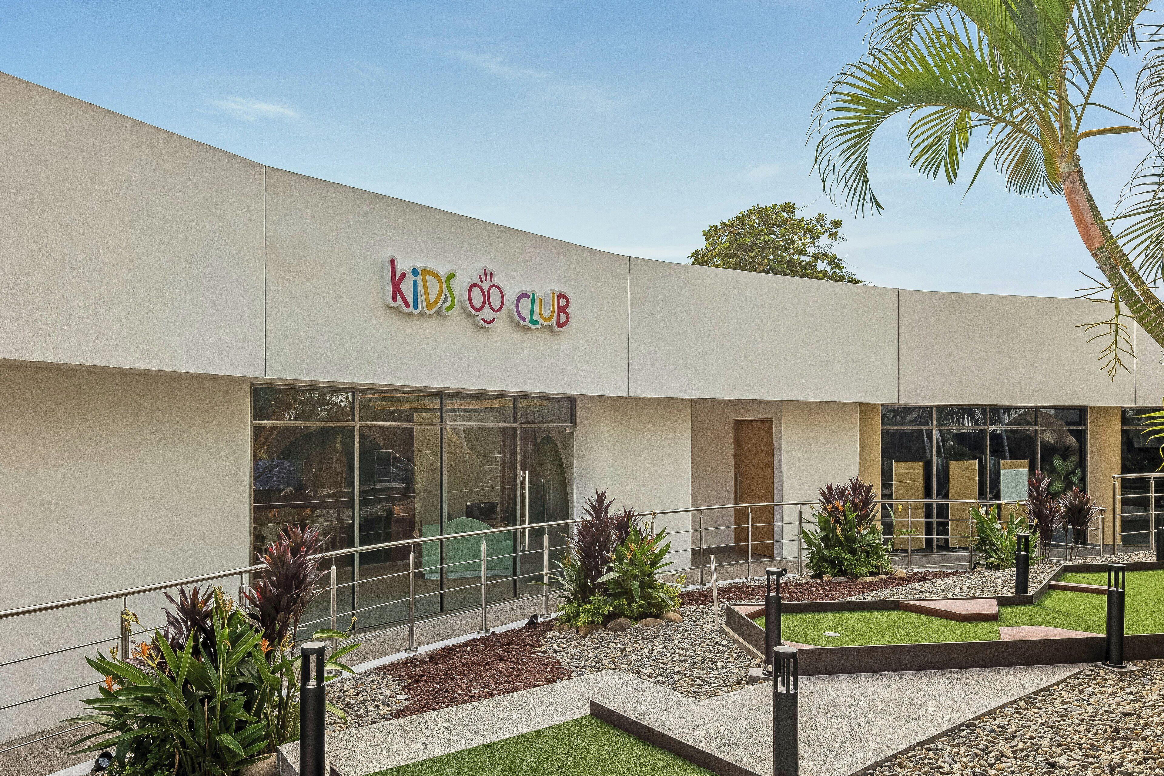 A building with a sign reading "Kids Club" and a landscaped outdoor area with plants and a palm tree.