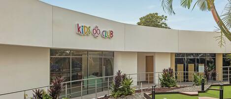 A building with a sign reading "Kids Club" and a landscaped outdoor area with plants and a palm tree.
