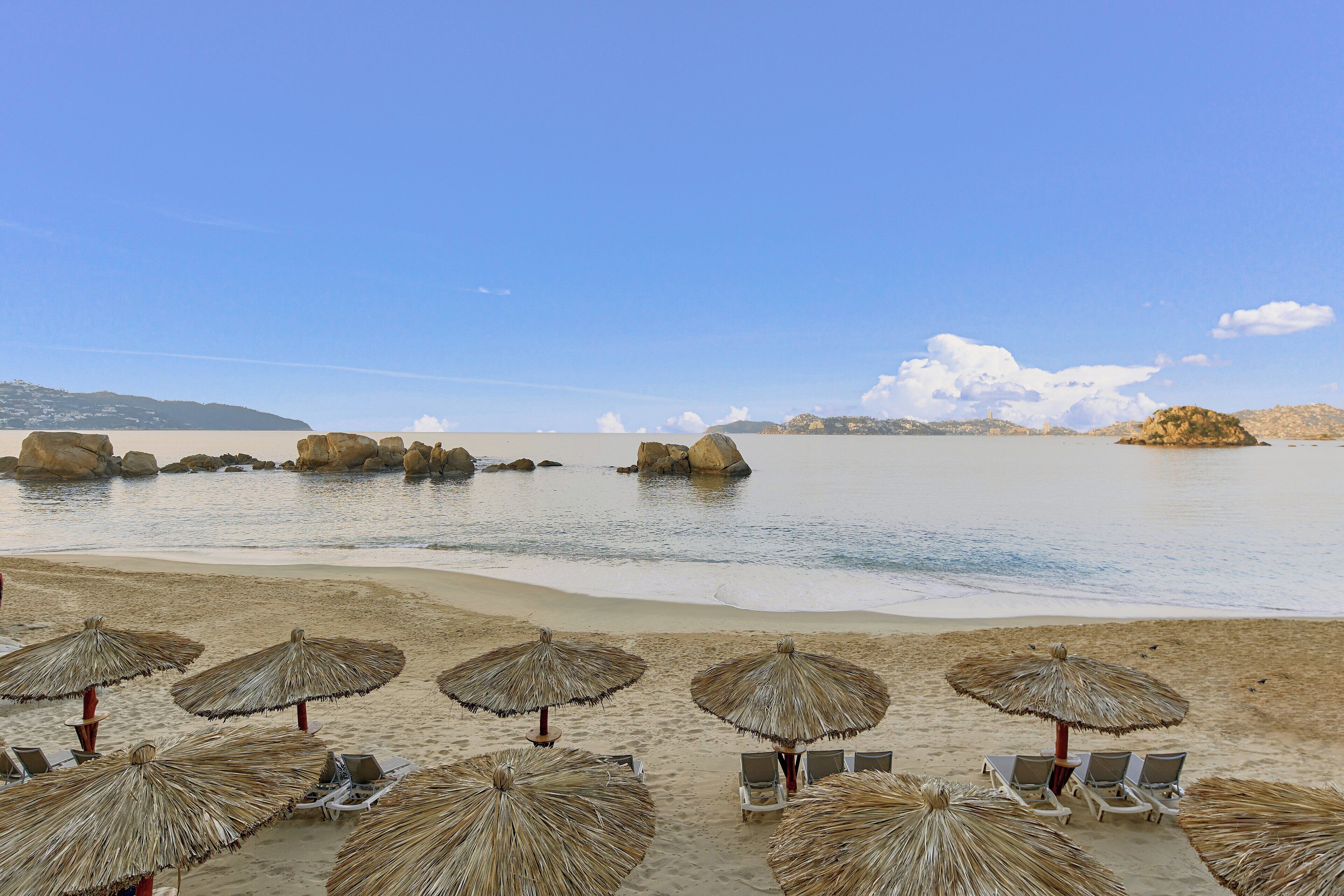 A beach with thatched umbrellas and lounge chairs, a calm sea, and rocky outcrops in the distance.