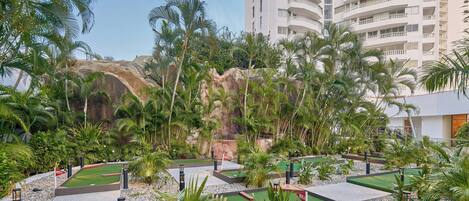 A mini-golf course with artificial turf, surrounded by palm trees and a rocky backdrop, adjacent to a high-rise building.