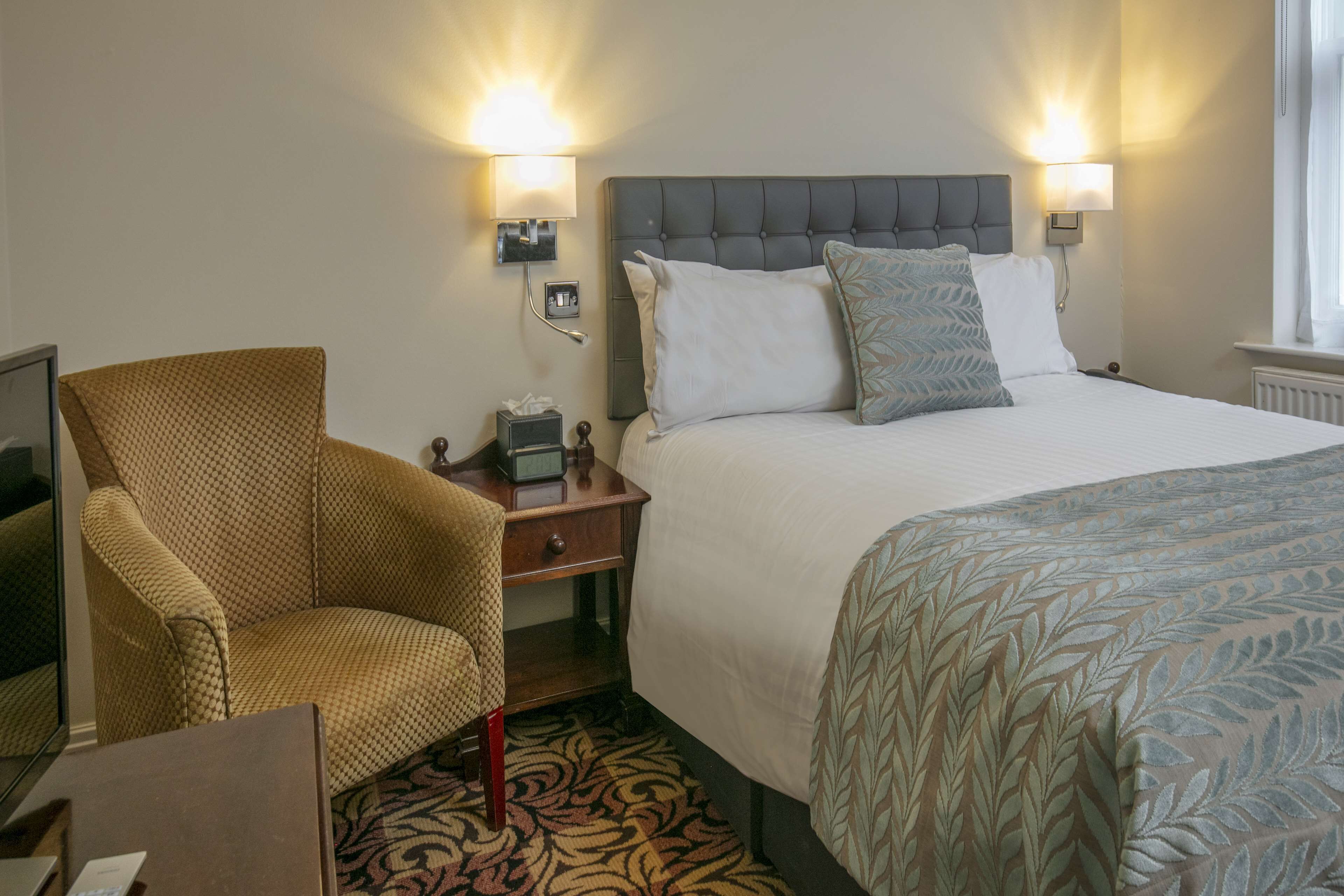 A hotel room with a bed, a chair, a nightstand, and two wall-mounted lamps.