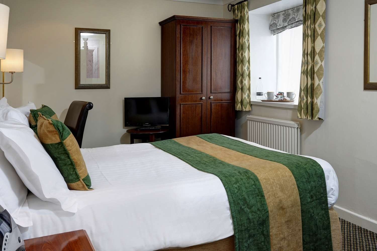 A hotel room with a bed, a television, a wardrobe, a window with curtains, and a radiator.