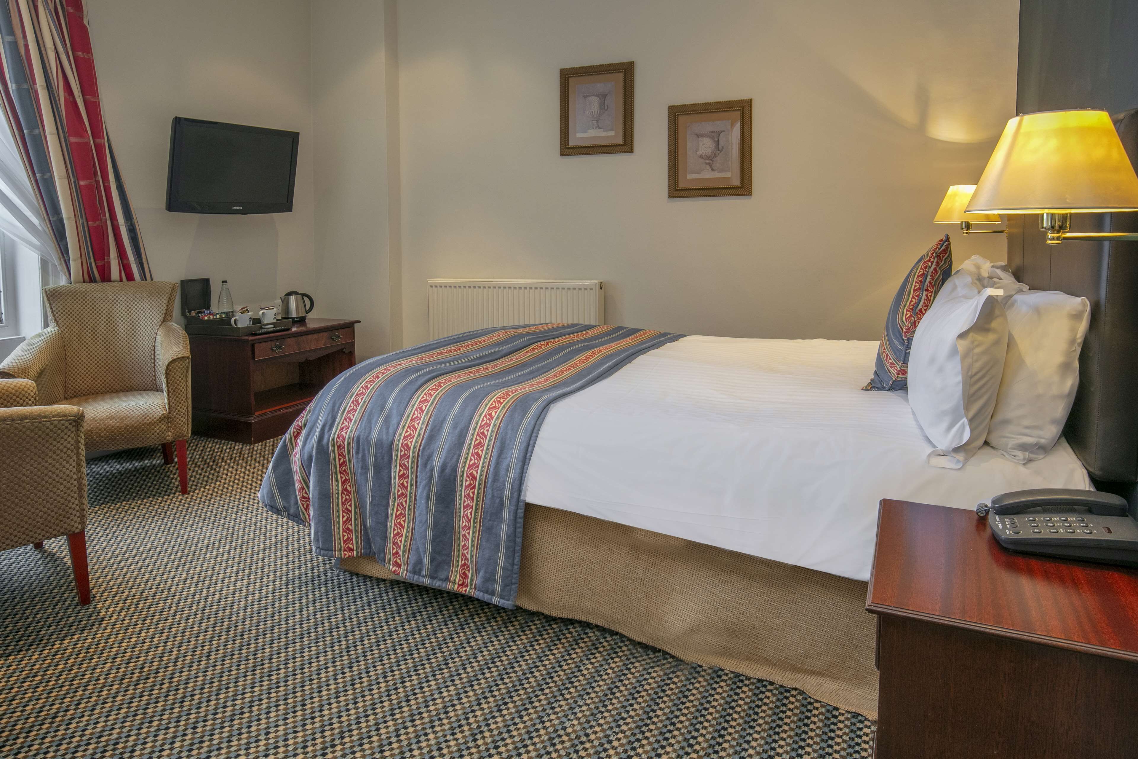 A hotel room with a bed, a chair, a desk with a lamp, a TV, and a window with curtains.