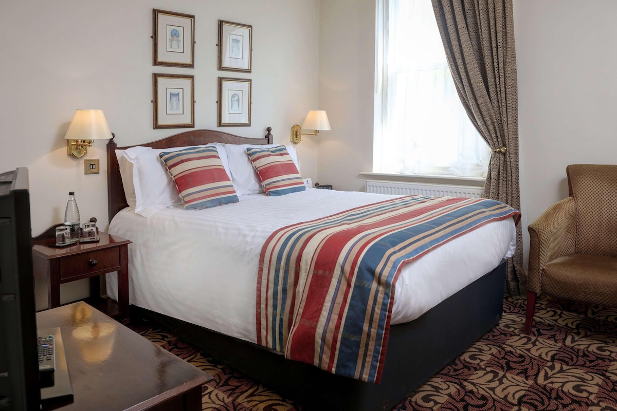 A hotel room with a bed, two bedside lamps, a television, a small table, and a chair.
