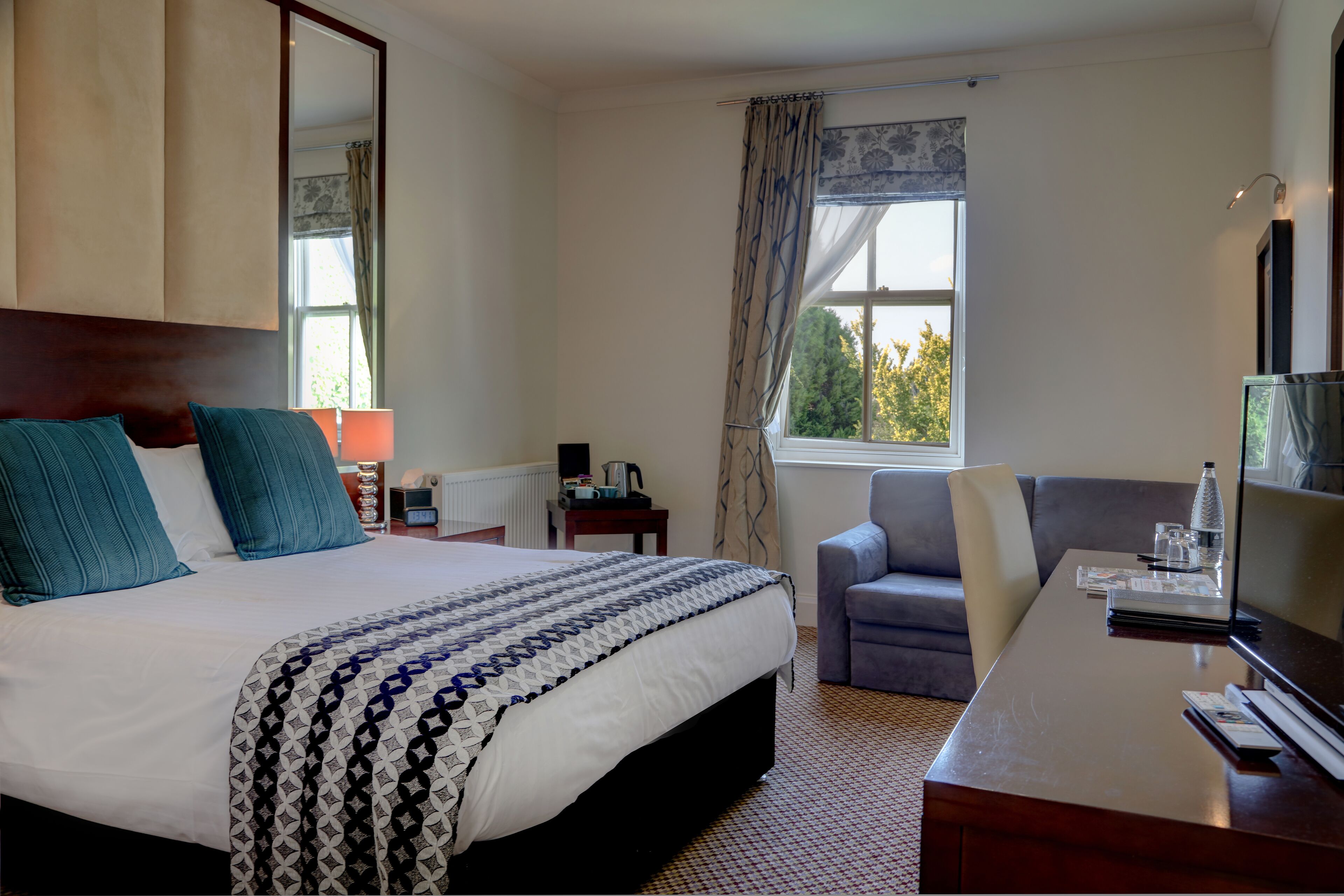 A hotel room with a large bed, a desk, a chair, and a window with a view of trees.