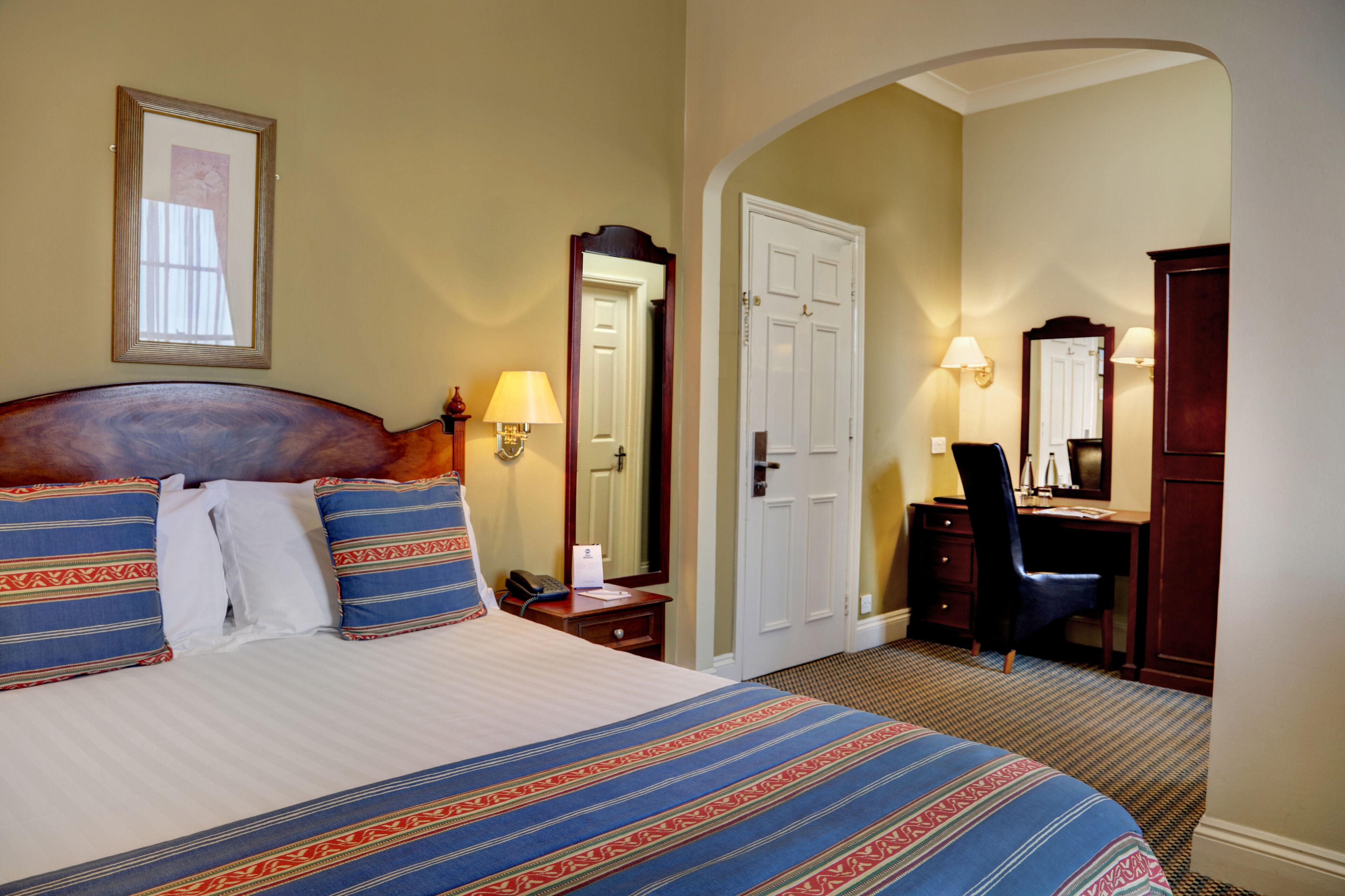 A hotel room with a large bed, a desk, a chair, a desk lamp, and a mirror.