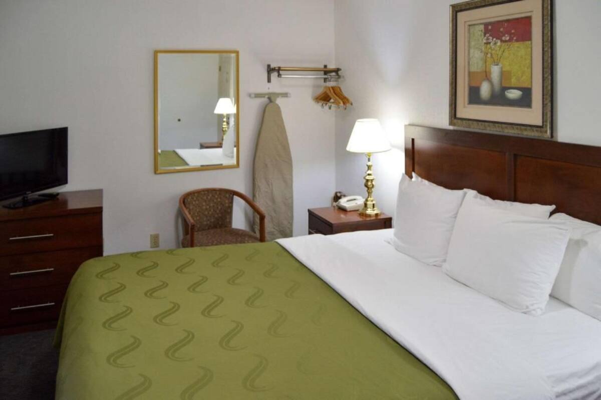 A hotel room with a bed, a television, a chair, a nightstand with a lamp, a mirror, and a painting on the wall.