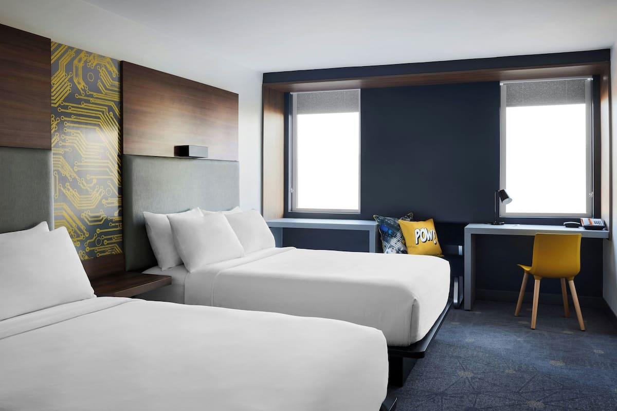 A hotel room with two beds, a desk, a chair, and a large window.