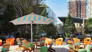Un espace extérieur aménagé avec des chaises Adirondack en bois et des chaises orange, un grand parasol rayé et une vue sur un paysage urbain avec de hauts immeubles.