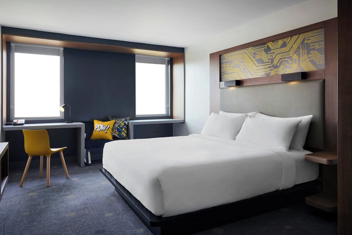 A hotel room with a large bed, a desk with a chair, and a window with blinds.